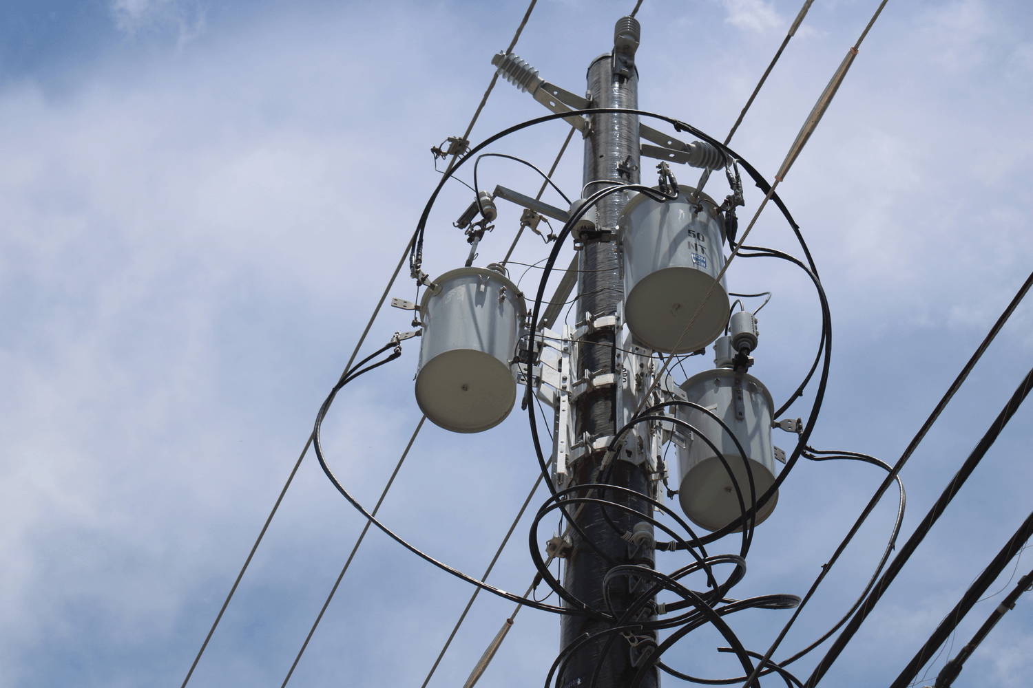 Utility pole with three cylindrical transformers and multiple tangled wires, set against a blue sky with light clouds.