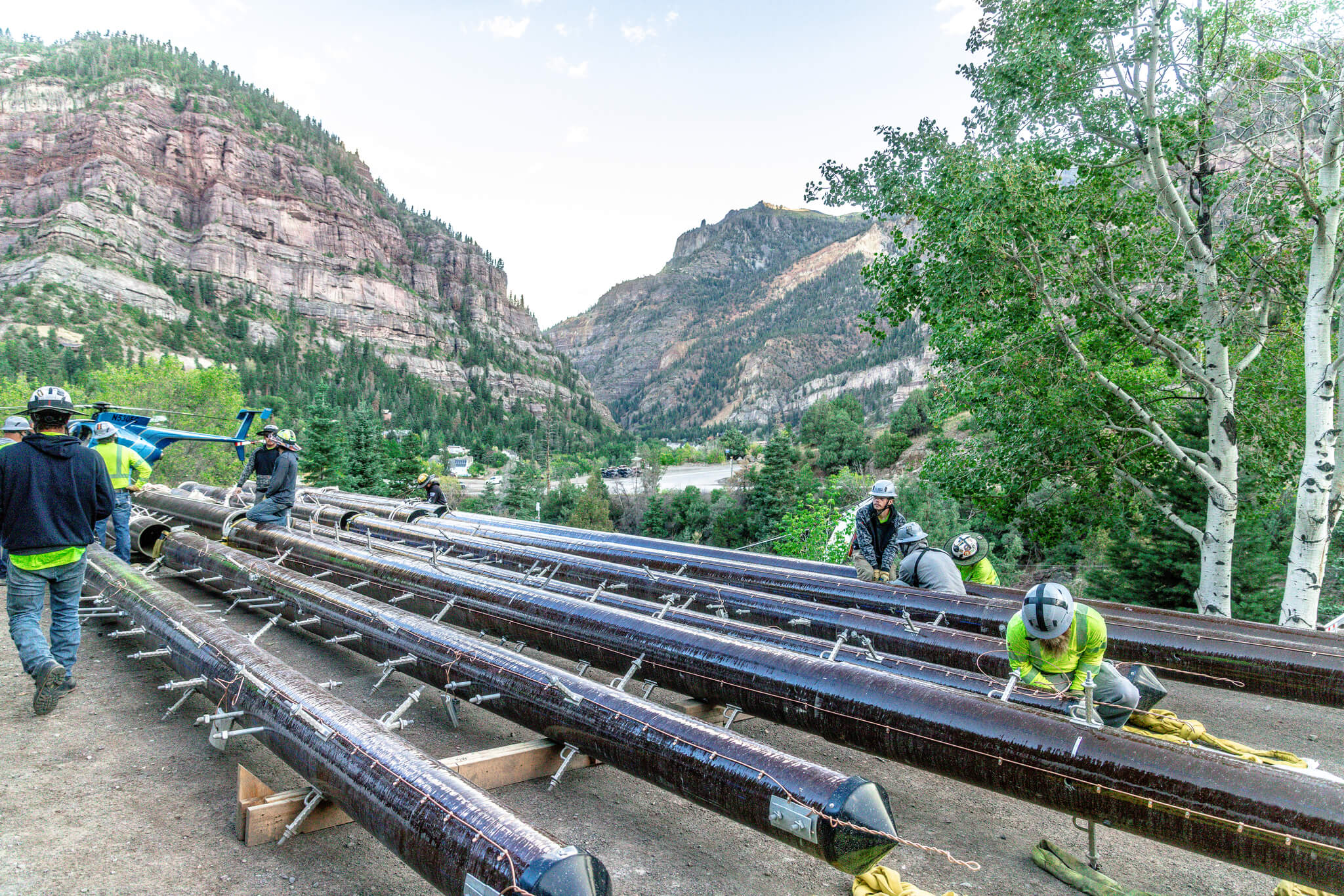 Workers in safety gear assemble or inspect large metal poles outdoors, surrounded by trees and rocky mountains under a clear sky.