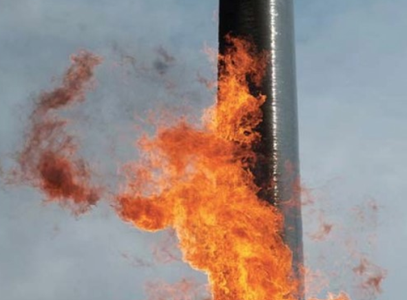 Bright orange flames are shooting from the side of a tall, cylindrical metal structure against a gray sky.