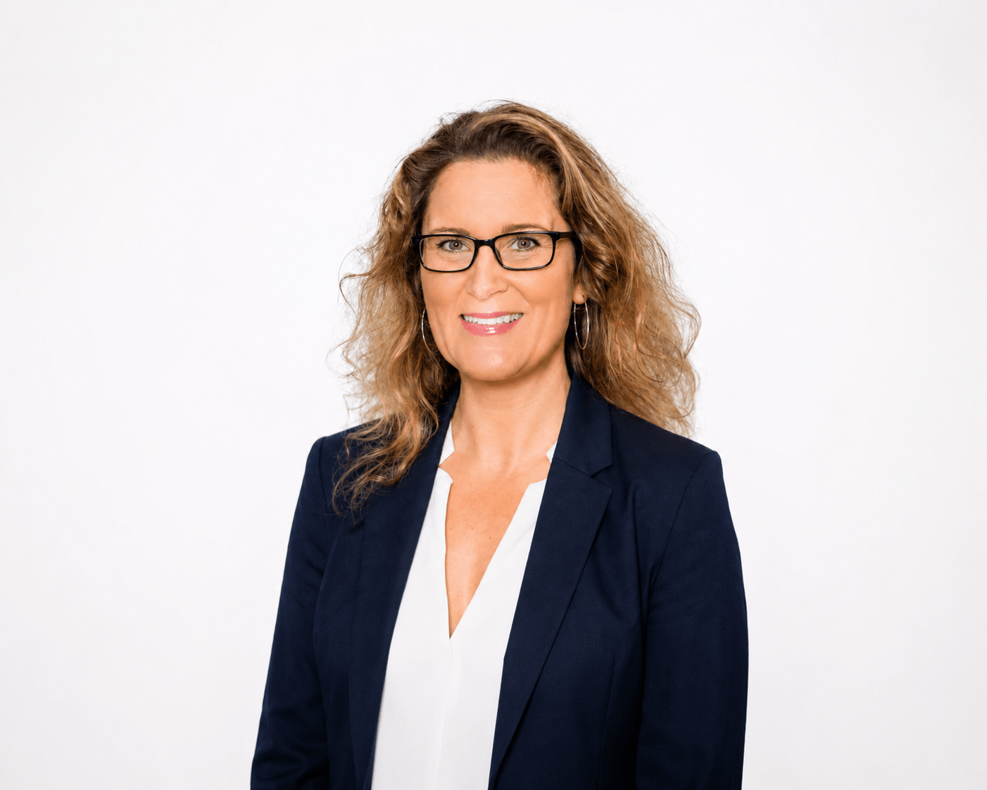 A woman with curly blonde hair and glasses smiles while wearing a navy blazer and white blouse, standing against a plain white background.