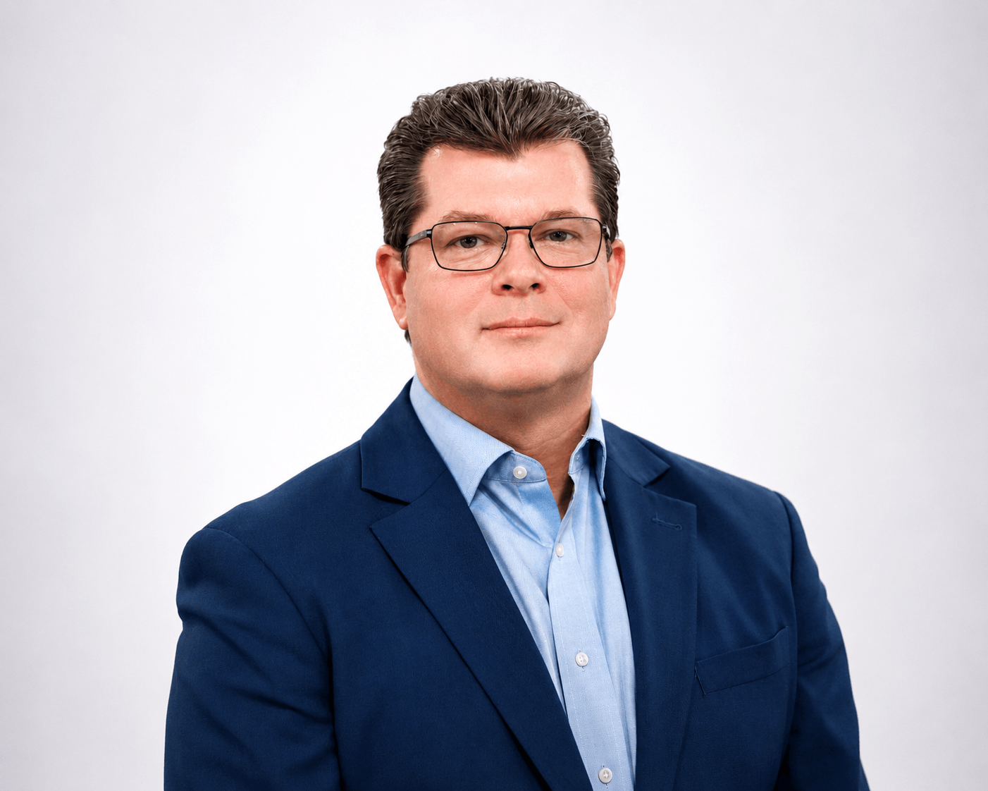 A man wearing glasses, a blue suit jacket, and a light blue shirt poses against a plain, light gray background, looking directly at the camera with a neutral expression.