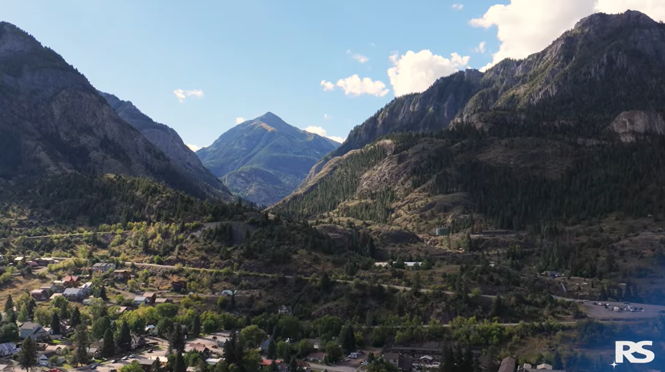 A scenic valley town surrounded by green trees and rugged mountains under a blue sky with scattered clouds. The letters RS appear in the bottom right corner.