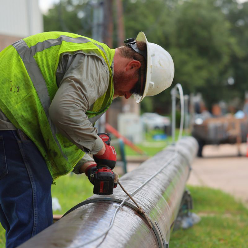 A worker in a safety vest and helmet uses a power drill on a large pipe outdoors, with trees and equipment visible in the background.