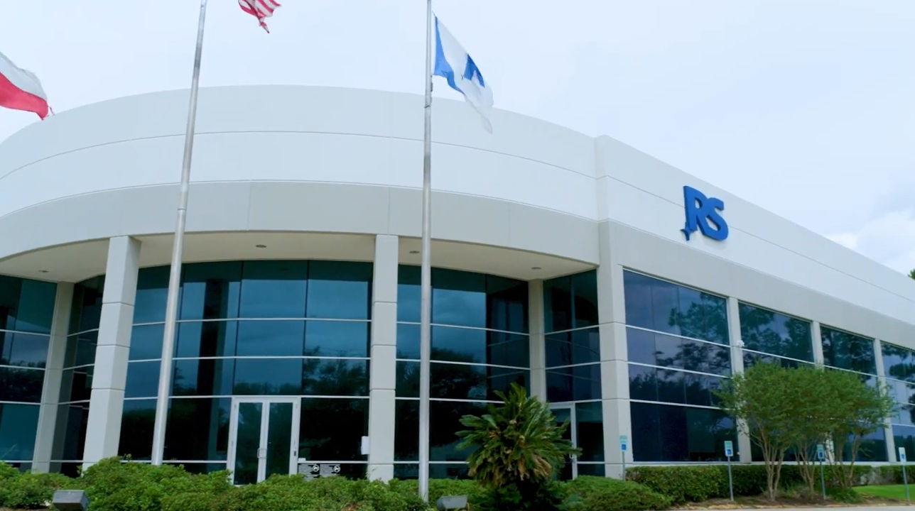 A modern white building with large glass windows, three flagpoles in front, and the blue RS logo on the upper right corner of the facade. Green landscaping lines the entrance.