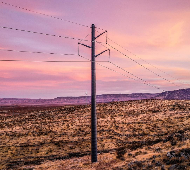 A power line pole stands in a dry, grassy landscape under a pink and purple sunset sky, with distant hills visible on the horizon.
