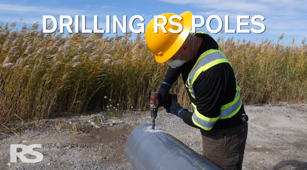 A worker in a hard hat and safety vest drills into a large gray pole outdoors, with tall grass in the background. Text reads DRILLING RS POLES..