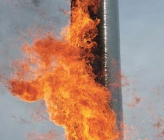 Bright orange flames engulf a vertical metal pipe or stack, set against a pale sky, suggesting a gas flare or industrial fire.
