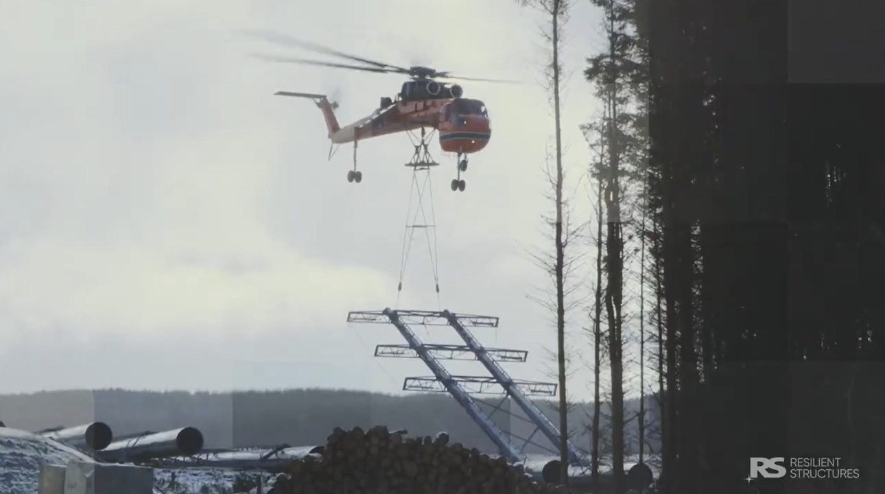 A large orange helicopter hovers above snowy ground, lowering a metal structure with cables between tall trees, in a forested area.