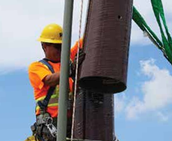 A construction worker in a yellow hard hat and orange vest secures a large cylindrical object to a pole against a blue sky.