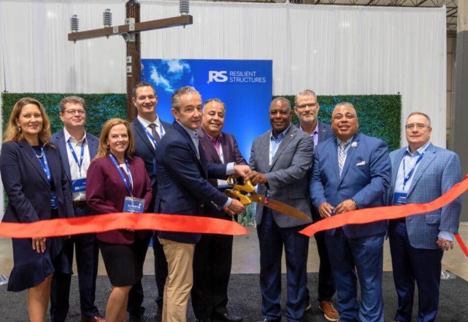 Ten people in business attire stand smiling behind a red ribbon, with two men in front cutting it with large scissors during a ribbon-cutting ceremony. A Resilient Structures banner is visible in the background.