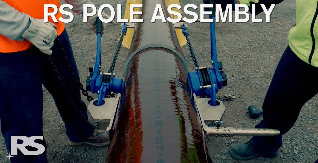 Two workers stand beside a large pole on the ground with blue equipment attached. Text reads RS POLE ASSEMBLY. The RS logo appears in the lower left corner.