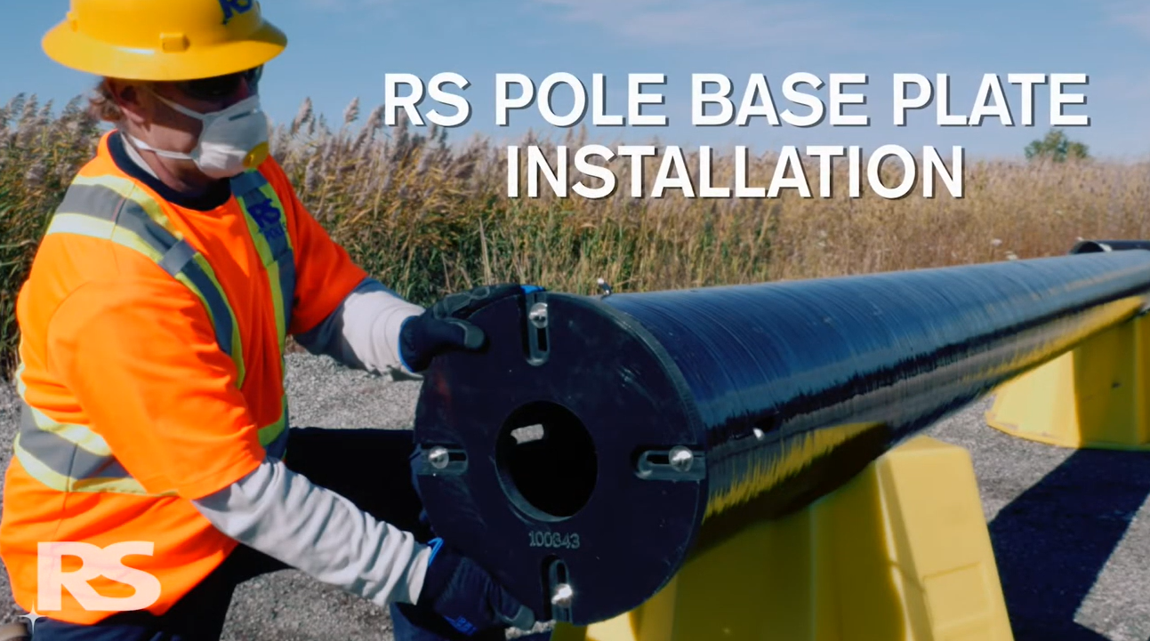 A worker wearing a hard hat and mask installs a large cylindrical pole onto yellow supports outdoors. Text over the image reads RS Pole Base Plate Installation..