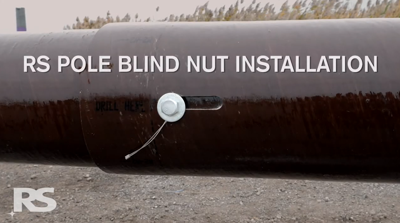 A large brown metal pole with a small silver nut and wire attached. The text RS POLE BLIND NUT INSTALL appears in white across the pole. Grassy field is blurred in the background.