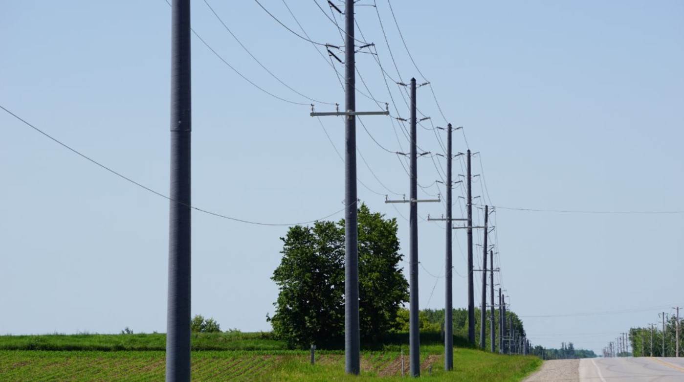 Utility poles with power lines run alongside a rural road bordered by green grass and a field, with a few trees and a blue sky in the background.