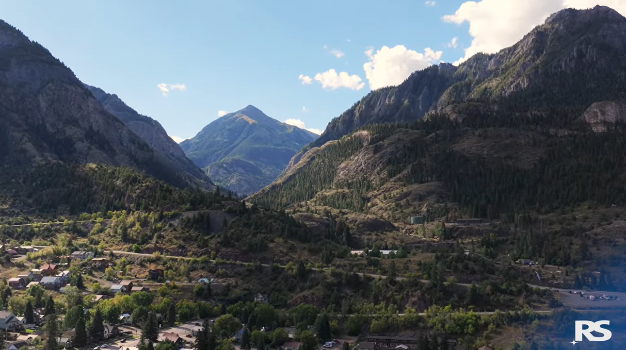 A scenic valley town surrounded by tall, green mountains under a bright blue sky with scattered clouds. Houses and roads are visible below.