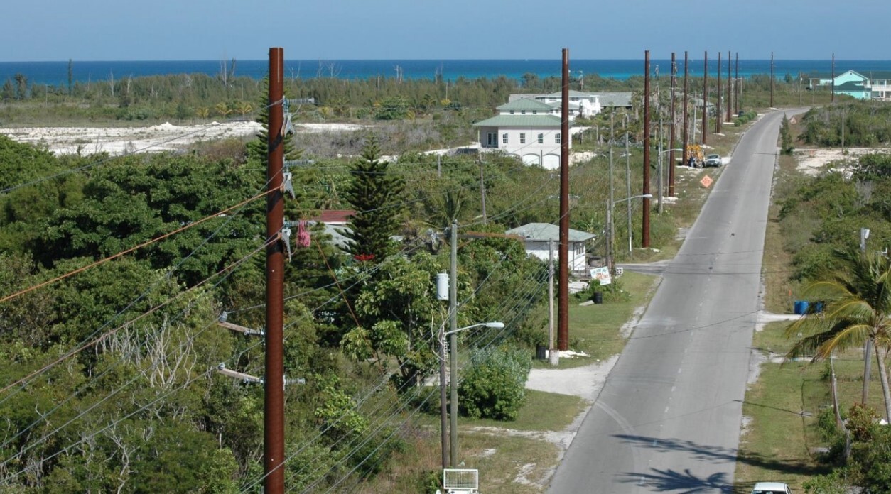 A paved road runs through a residential area with utility poles on both sides, surrounded by green trees and houses, leading toward the ocean under a clear blue sky.