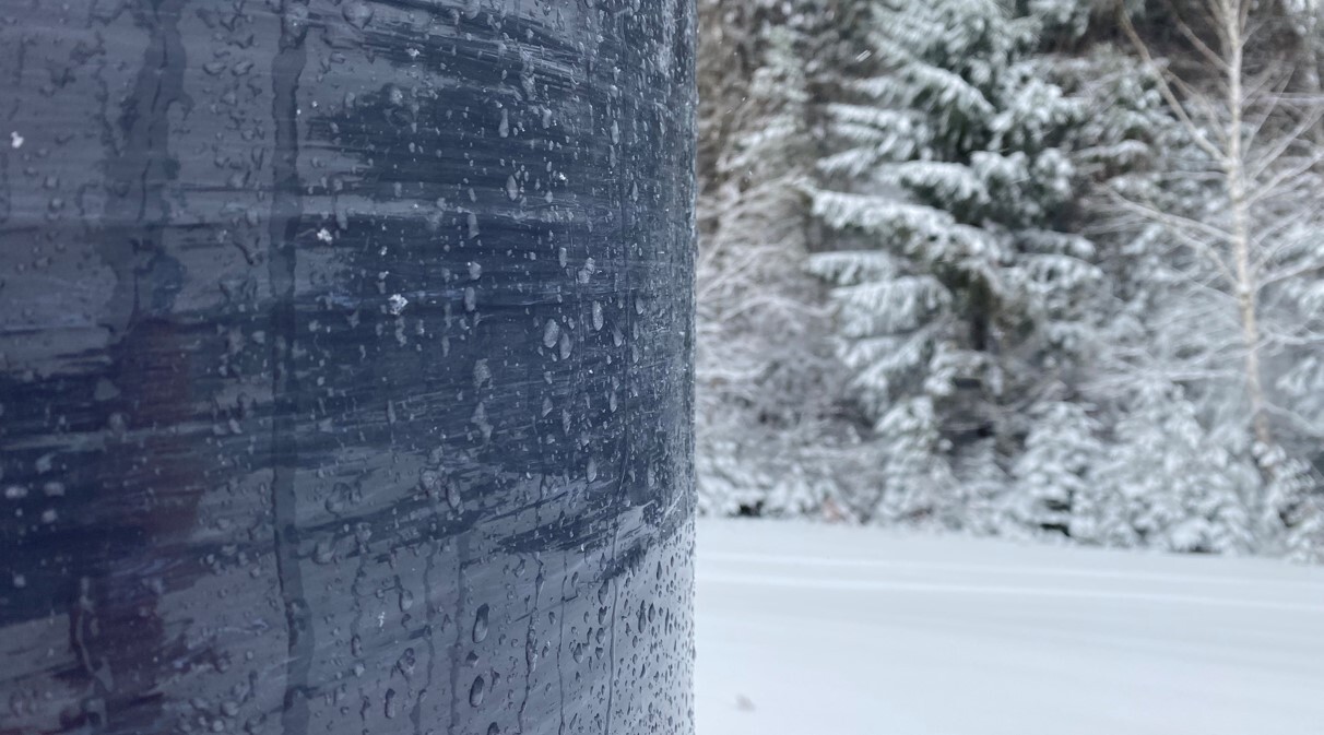 Close-up of a wet, dark pole with water droplets, set against a snowy landscape with trees covered in snow in the background.